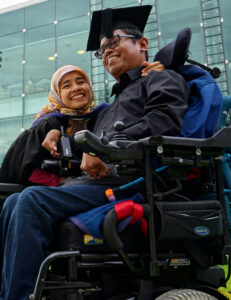 Faisal Rusdi photograph - Faisal is to the right and his wife, Cucu, to the left. They are in wheelchairs and sitting on the green lawn. Behind them is the University Building